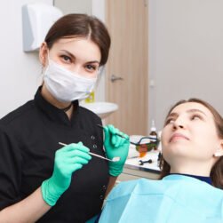 Indoor shot of skillful confident young female dentist wearing exam gloves and white mask holding metal probe and dental miror, ready to examine oral cavity of woman patient sitting in chair